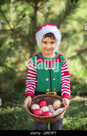 Half-length portrait of a little boy in elf hat and red sweater eating cookie near christmas tree outside waiting for a Christmas in the wood. Active outdoors leisure with children Stock Photo