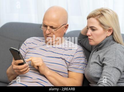 Smiling older father with adult daughter using phone together, having fun, watching video in social network or shopping online, caring young woman Stock Photo