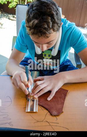 A teenager uses a sharp scalpel and metal ruler as a guide to cut a straight piece of leather. Stock Photo
