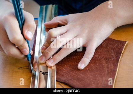 Close up view of a teenager's hands using a sharp scalpel and metal ruler as a guide to cut a straight piece of leather. Stock Photo