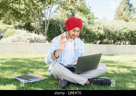 Cheerful indian male student greeting participants of virtual meeting, hindu man in turban sits with laptop and waving hello into webcam, has video conference with classmates sitting outdoors Stock Photo