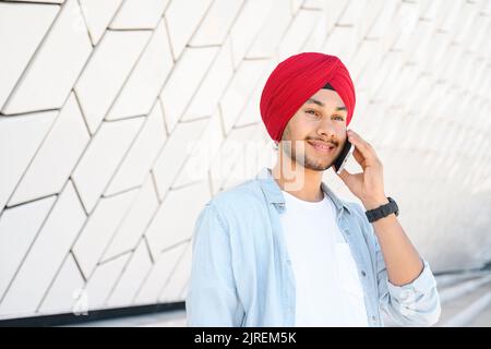 Intelligent indian male office employee, freelancer or student talking on the smartphone outdoors, eastern man in traditional red headwear turban has pleasant phone conversation Stock Photo