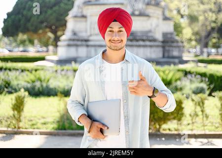 Positive smiling Indian man freelancer in turban showing thumbs up standing outdoors and holding laptop, satisfied hindu male student recommends studying on the distance from anywhere in the world Stock Photo