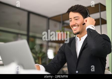 Portrait of confident latin man in wireless headset using laptop computer sitting in contemporary office space, male employee is making video call, talking online on distance Stock Photo