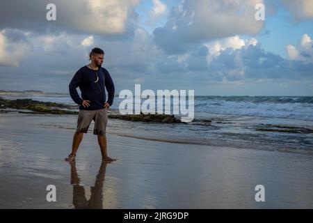 Middle-aged bearded man on the beach during sunrise looking lost at the landscape. Stock Photo