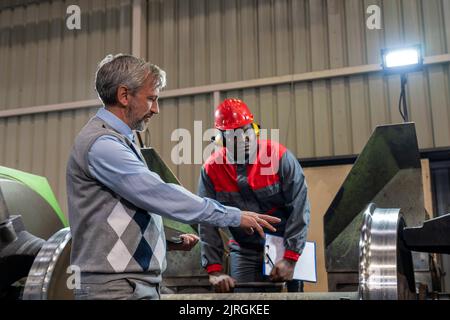Quality Control Inspector Talking To African American CNC Operator Next To Lathe Machine In A Train Factory. Train Wheel Manufacturing. Stock Photo