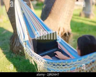 Young business woman working in a hammock. Young lady downshifter working at laptop and enjoys and relaxed environment, working day. Online freelance work on vacation. Stock Photo