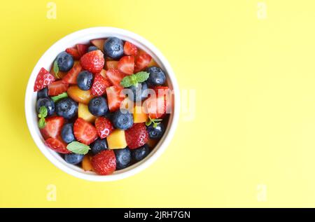 Fruit salad in a white cup on a yellow background. Sliced strawberries, peaches and blueberries with mint leaves. Free space for text. Stock Photo