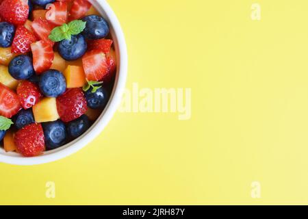 Fruit salad in a white cup on a yellow background. Sliced strawberries, peaches and blueberries with mint leaves. Free space for text. Stock Photo