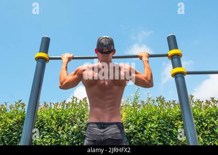 Muscular man in a cap doing pull-ups on the horizontal bar in the park outdoors, back view of his back Stock Photo
