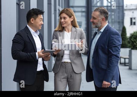 Three businessmen outside office building with documents discussing plans, businessmen and businesswoman having fun chatting and talking, diverse business group in business suits Stock Photo