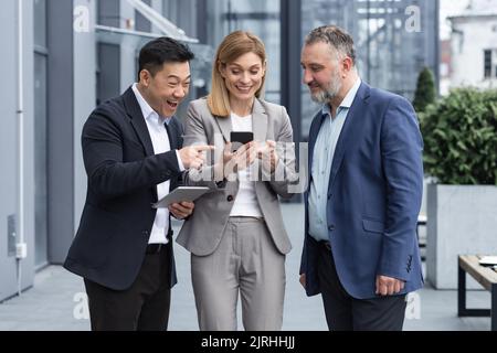 Three colleagues employees watch funny videos or look at photos, pictures memories looking at smartphone screen using mobile phone. group of business people office workers having fun on breaks Stock Photo