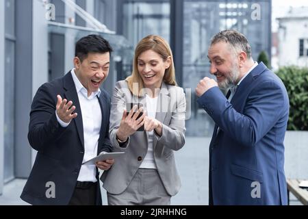 Three colleagues employees watch funny videos or look at photos, pictures memories looking at smartphone screen using mobile phone. group of business people office workers having fun on breaks Stock Photo