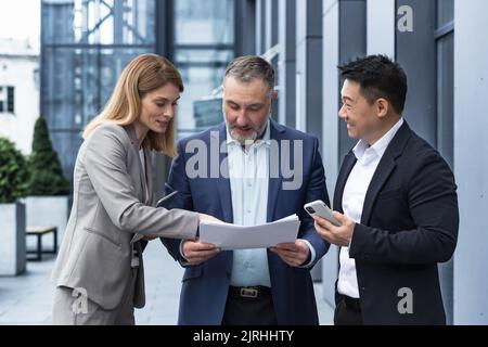 Three businessmen outside office building with documents discussing plans, businessmen and businesswoman having fun chatting and talking, diverse business group in business suits Stock Photo