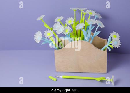Blue-green daisies-shaped pens in a wooden box on a table on a purple background. Back to school Stock Photo