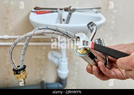 Plumbing work on installing water faucet on sink in bathroom, plumber fixes braided connecting hoses on tap using adjustable wrench. Stock Photo