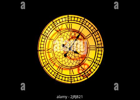 Glowing vintage yellow clock with roman dial on dark wall. Blurred image. Stock Photo