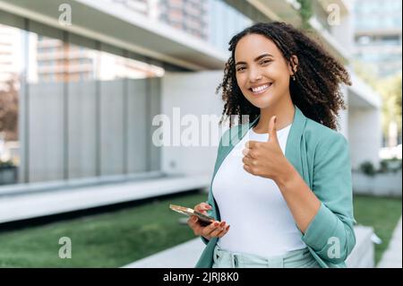 Beautiful positive confident hispanic or brazilian curly young woman, in stylish clothes, walks down the street, holds her smartphone in her hand, looks at the camera, smiles, shows thumbs up gesture Stock Photo