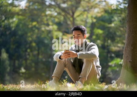 young asian adult man sitting on grass in park looking at cellphone Stock Photo