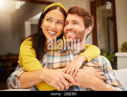 Happy and excited couple hugging thinking of future relaxing and sitting on a couch at home. Relaxed diverse interracial lovers smiling and enjoying Stock Photo