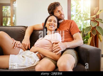 Happy, love and interracial couple smile looking away relax and sitting on a couch at home. Diversity man and woman smiling and enjoying quality Stock Photo