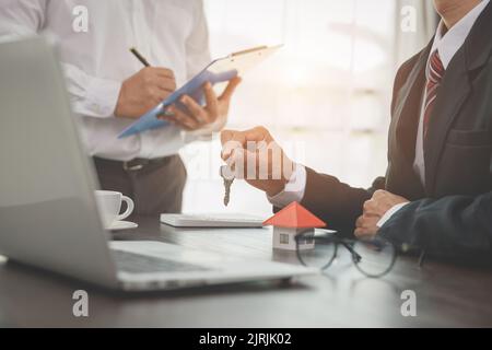 Businessman signs contract behind home architectural model, female hand reviewing real estate contract. Stock Photo