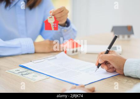 A woman signs a contract to purchase a home with a real estate agent. Model house with keys on table. Stock Photo