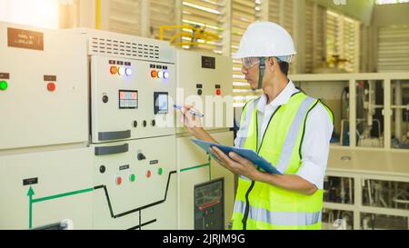 A helmeted technician holds a clipboard while checking and taking notes on the electrical control cabinet. Stock Photo