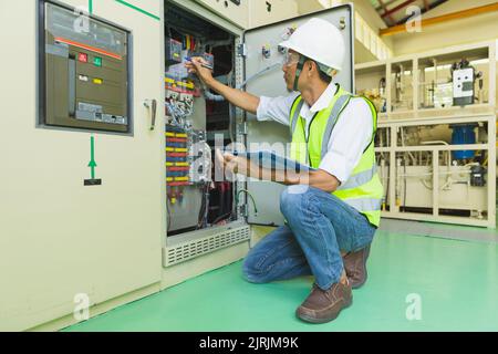 A helmeted technician holds a clipboard while checking and taking notes on the electrical control cabinet. Stock Photo