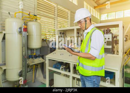 A helmeted technician holds a clipboard while checking and taking notes on the electrical control cabinet. Stock Photo
