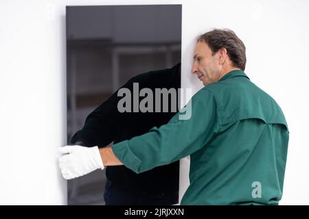 Tauberbischofsheim, Germany. 18th Aug, 2022. An employee of the manufacturer of infrared heaters Vitramo attaches an infrared heater for wall mounting to the wall. In an infrared heater, a frontal cover (glass or sheet metal) is heated from the back by a heating layer that converts electrical energy into heat. Credit: Daniel Karmann/dpa/Alamy Live News Stock Photo