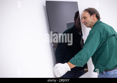 Tauberbischofsheim, Germany. 18th Aug, 2022. An employee of the manufacturer of infrared heaters Vitramo attaches an infrared heater for wall mounting to the wall. In an infrared heater, a frontal cover (glass or sheet metal) is heated from the back by a heating layer that converts electrical energy into heat. Credit: Daniel Karmann/dpa/Alamy Live News Stock Photo