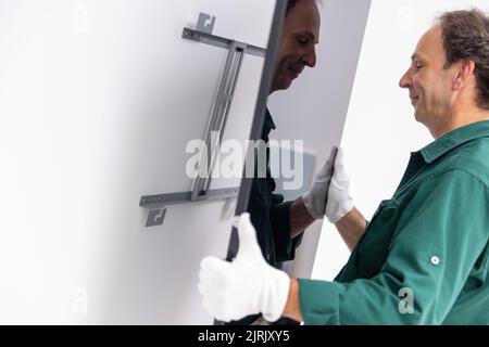 Tauberbischofsheim, Germany. 18th Aug, 2022. An employee of the manufacturer of infrared heaters Vitramo attaches an infrared heater for wall mounting to the wall. In an infrared heater, a frontal cover (glass or sheet metal) is heated from the back by a heating layer that converts electrical energy into heat. Credit: Daniel Karmann/dpa/Alamy Live News Stock Photo