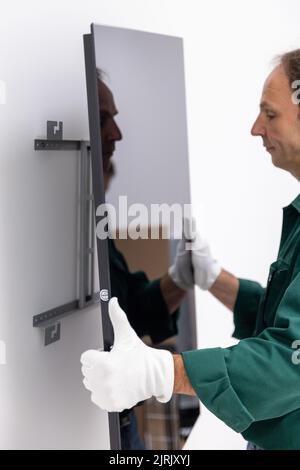 Tauberbischofsheim, Germany. 18th Aug, 2022. An employee of the manufacturer of infrared heaters Vitramo attaches an infrared heater for wall mounting to the wall. In an infrared heater, a frontal cover (glass or sheet metal) is heated from the back by a heating layer that converts electrical energy into heat. Credit: Daniel Karmann/dpa/Alamy Live News Stock Photo