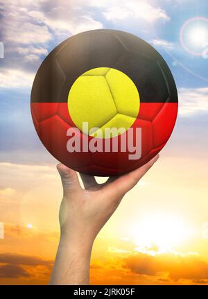 Soccer ball in hand with a depiction of the flag of Australian Aboriginal against a colorful sky Stock Photo