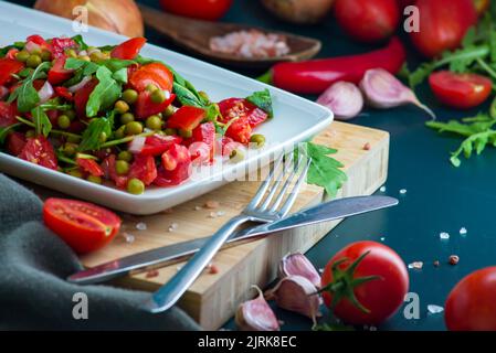 summer salad, tomatoes, green peas, arugula, onion, olive oil, garlic, hot pepper fork knife bamboo cutting board pastel green napkin  textile, salt V Stock Photo