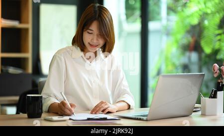 Pretty young woman office worker using laptop computer and preparing annual financial report at office desk Stock Photo