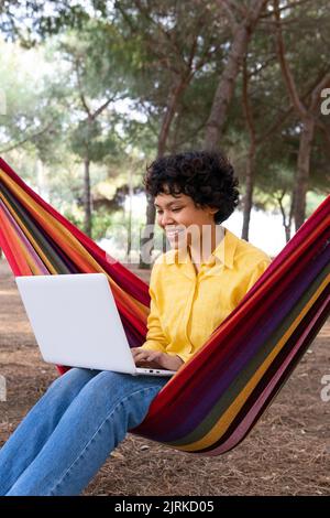 Smiling teen female chilling in hammock in park and browsing laptop Stock Photo