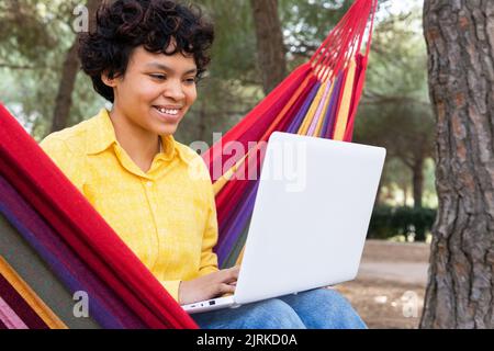Smiling teen female chilling in hammock in park and browsing laptop Stock Photo