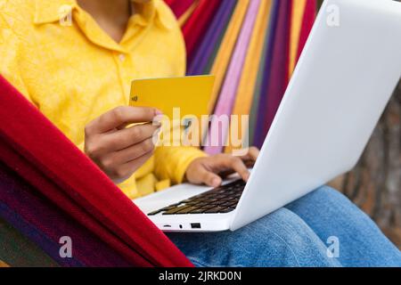 Smiling young female sitting in hammock in park and using laptop while holding credit card buying online Stock Photo