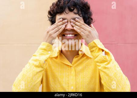 Content young ethnic female with short dark curly hair wearing vivid yellow shirt and closing eyes with hands Stock Photo