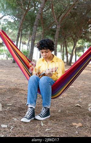 Smiling young ethnic female in bright shirt lounging in park hammock and playing ukulele Stock Photo