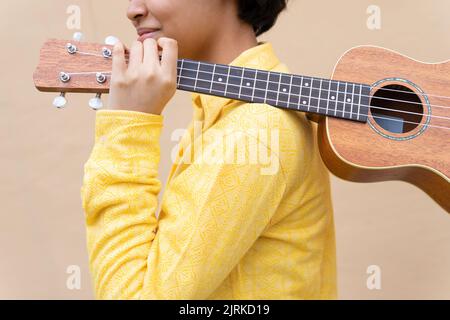 Side view of cropped unrecognizable charming ethnic female with short curly hair holding ukulele guitar over shoulder standing near colorful backgroun Stock Photo