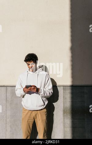 Young smiling male standing on concrete wall in the city street and browsing mobile phone Stock Photo