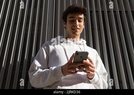 From below of young content male standing near metal wall in city and messaging on social media via mobile phone Stock Photo
