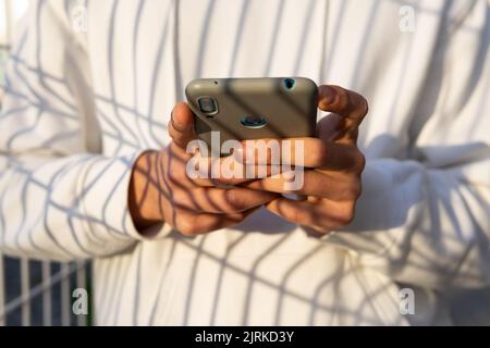 Cropped unrecognizable male surfing Internet on mobile phone while standing near metal net and looking away Stock Photo
