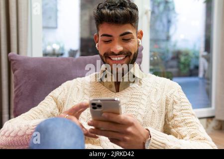 Smiling young Hispanic male with beard and dark hair sitting at home and browsing mobile phone Stock Photo