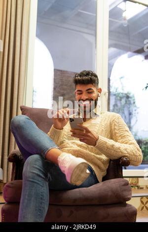 Smiling young Hispanic male with beard and dark hair sitting at home and browsing mobile phone Stock Photo