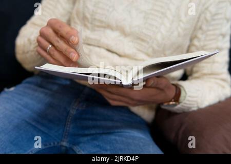 Crop of anonymous young Hispanic male in stylish outfit lying on comfortable sofa while reading interesting book Stock Photo