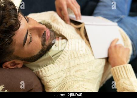 Top view of young Hispanic bearded male in stylish outfit lying on comfortable sofa and sleeping after reading interesting book Stock Photo